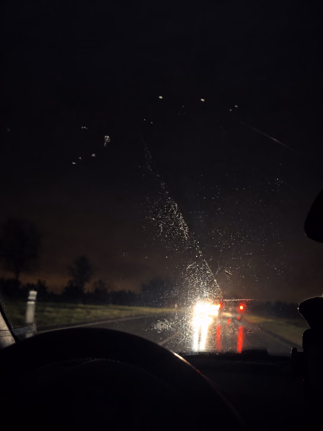 Blick durch eine nasse Frontscheibe bei Nacht, Regentropfen auf dem Glas und Lichter der Autos vor dir