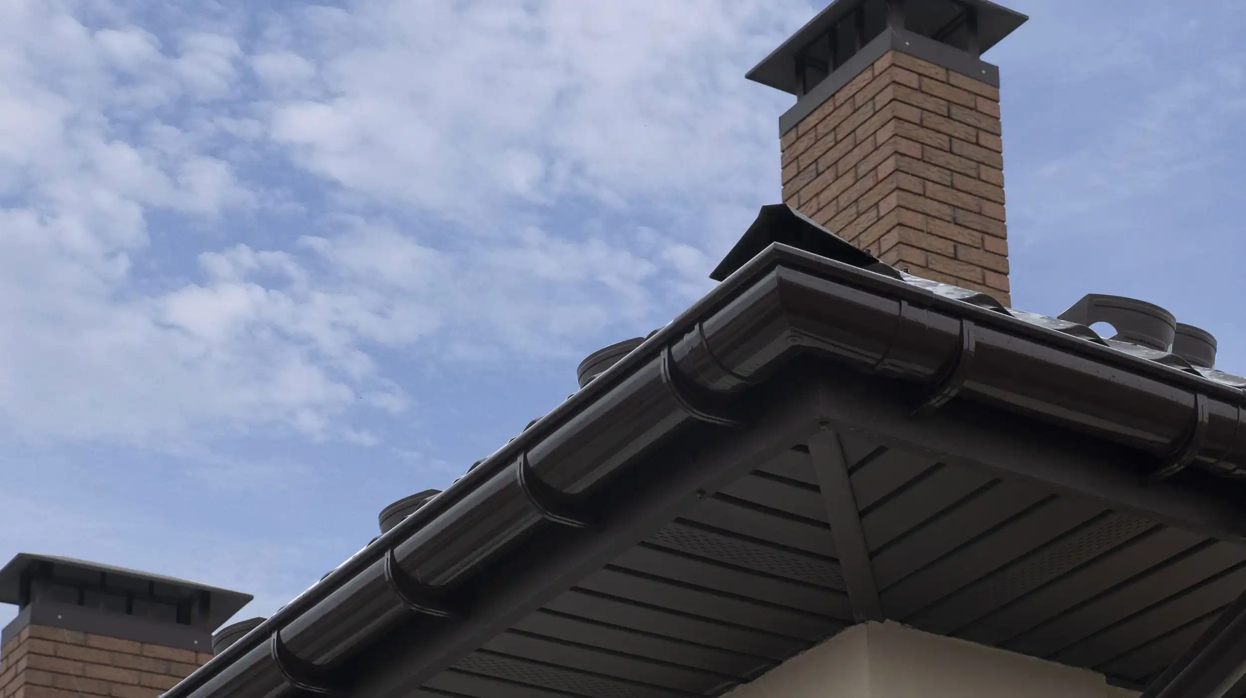 Roof corner with black guttering, fascia boards, and brick chimney against a partly cloudy sky