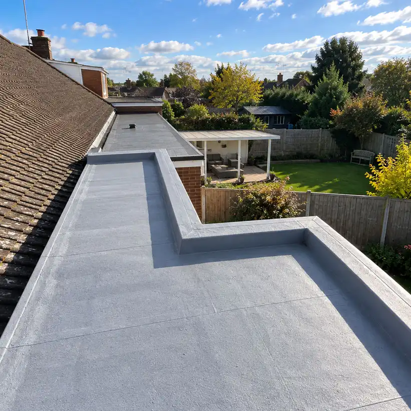 Opposite angle view of a flat roof extension with grey waterproof membrane, showing parapet edges, tiled roof to the left, and rear garden with fencing and outbuildings under a bright sky