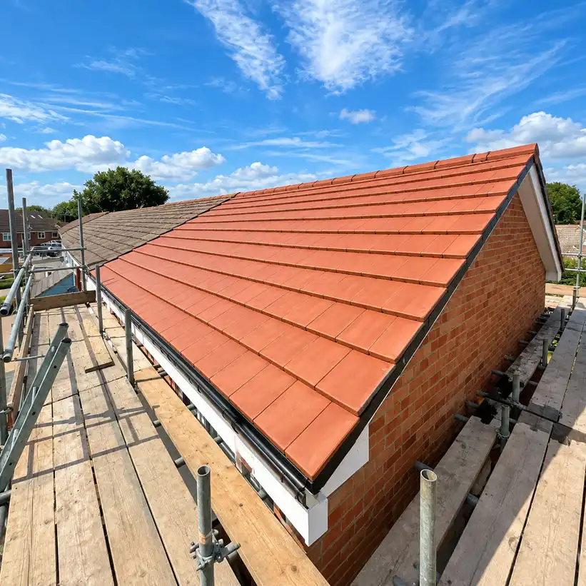 Wide-angle view of a newly installed red-orange tiled pitched roof on a brick house, surrounded by scaffolding and wooden platforms under a bright blue sky with scattered clouds