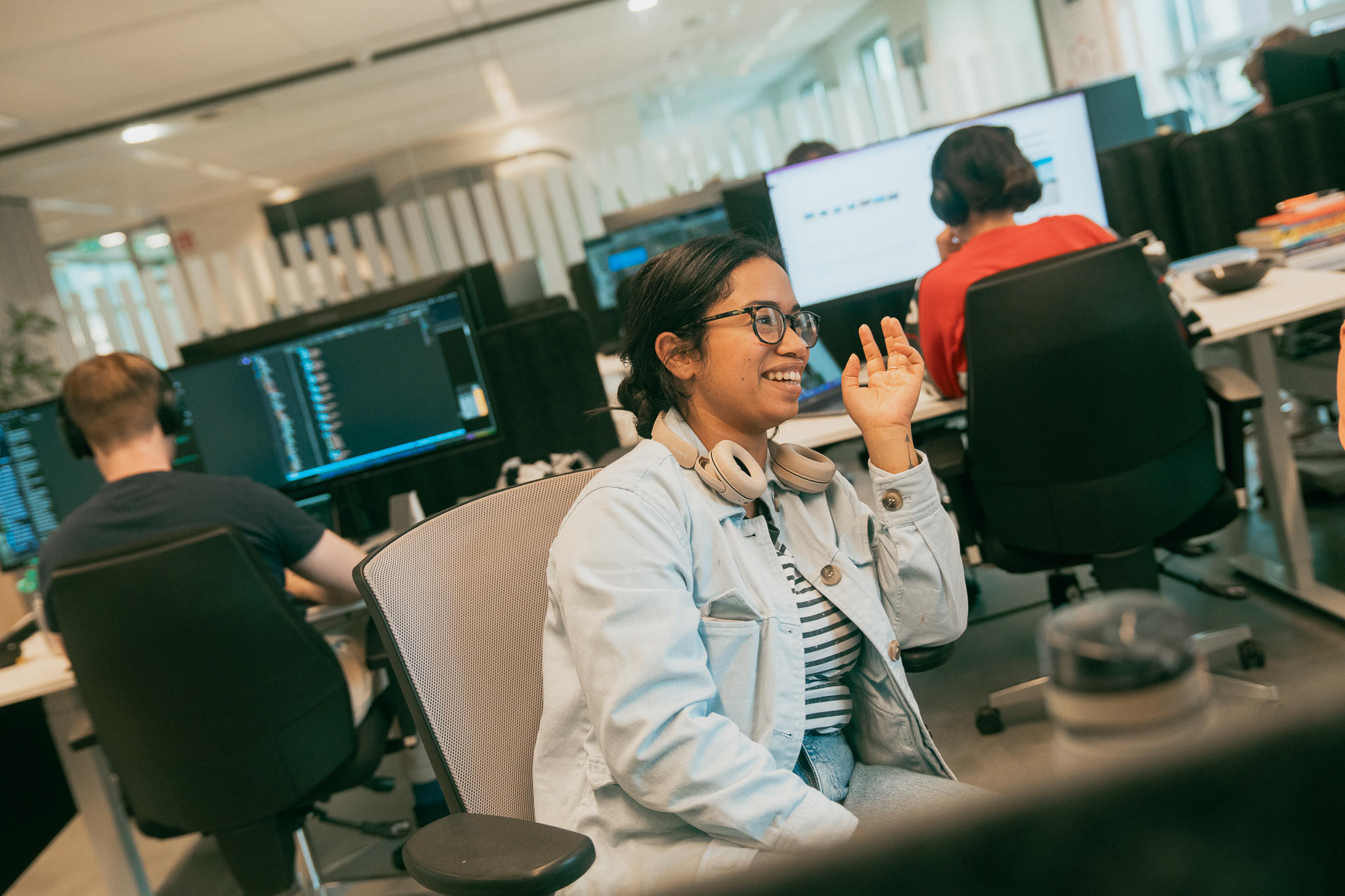 Een glimlachende vrouw met een koptelefoon om haar nek, die in een kantoor met collega’s achter computers in gesprek is.