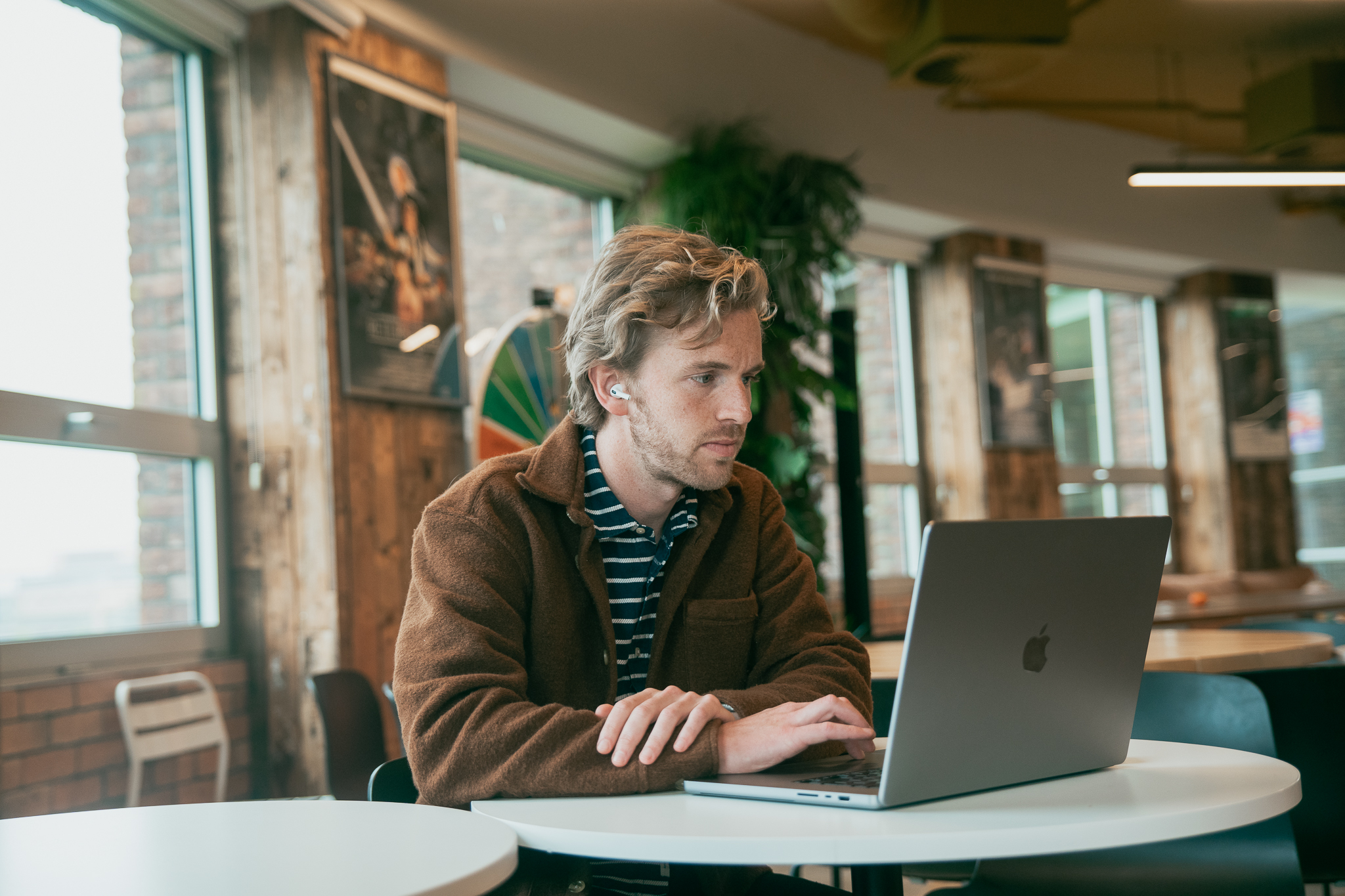 Een man met oortjes die geconcentreerd naar zijn laptop kijkt in een moderne werkruimte met houten wanden en grote ramen.