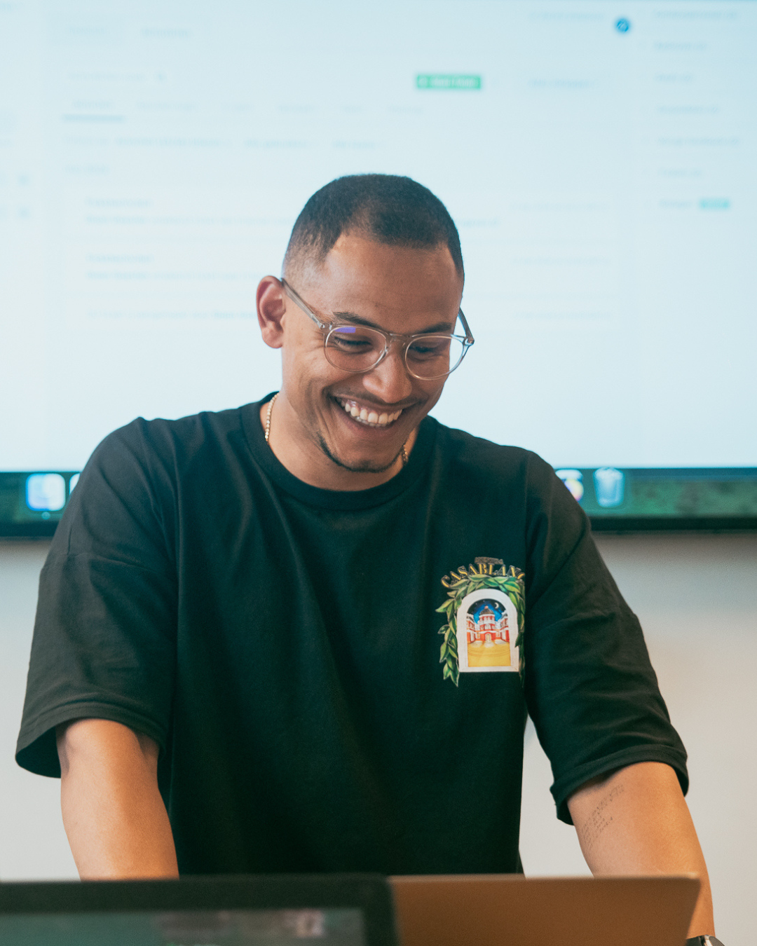 Een glimlachende man met een bril en een zwart Casablanca-shirt, die op een laptop werkt, met een scherm op de achtergrond.