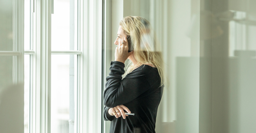 Woman with blonde hair wearing a black long-sleeve shirt, standing by a window, talking on a smartphone while holding a pen.