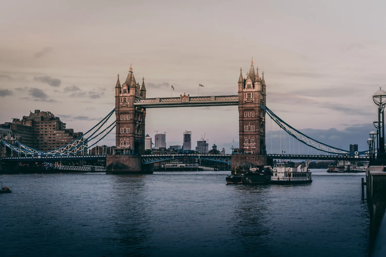 Evening view of the Tower Bridge over the River Thames in London with boats near the bridge and city skyline in the background under a cloudy sky.
