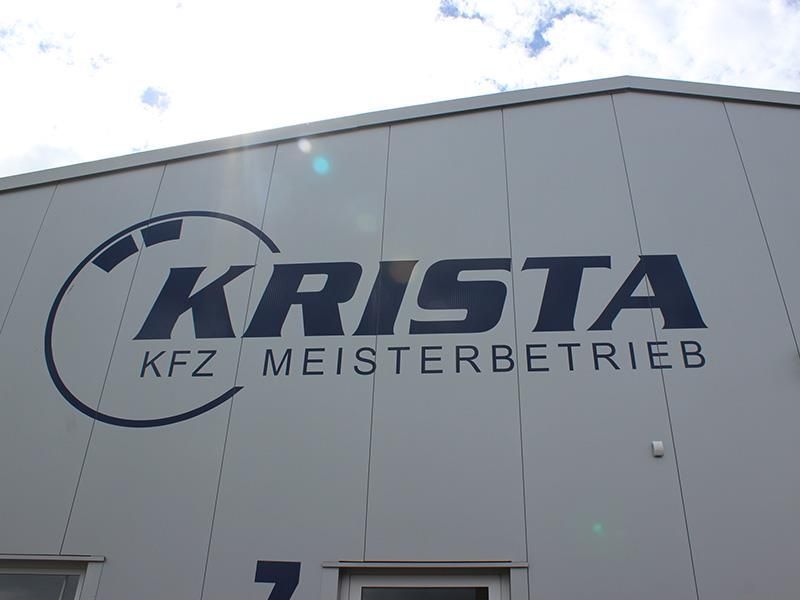 Logo und Schriftzug der Firma KRISTA KFZ Meisterbetrieb an der Fassade eines hellen Industriegebäudes unter bewölktem Himmel.