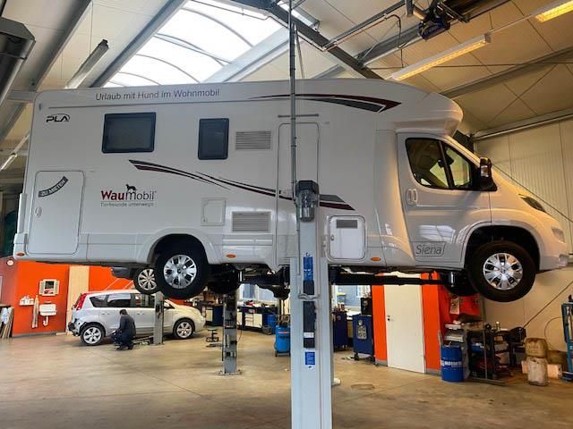 Weißes Wohnmobil mit der Aufschrift "Urlaub mit Hund im Wohnmobil" und "Waumobil" auf der Seite steht auf einer Hebebühne in der KFZ-Werkstatt Krista, im Hintergrund ist ein weiteres Fahrzeug mit einer Person zu sehen.