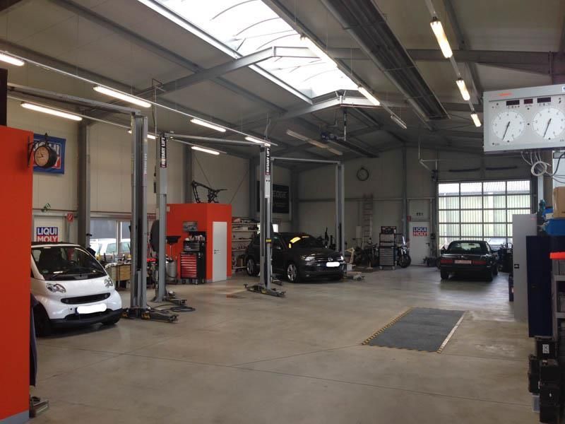 Innenansicht einer großen, gut beleuchteten Autowerkstatt mit mehreren Hebebühnen, drei geparkten Autos und orangefarbenen Wänden an den Werkstattstationen.