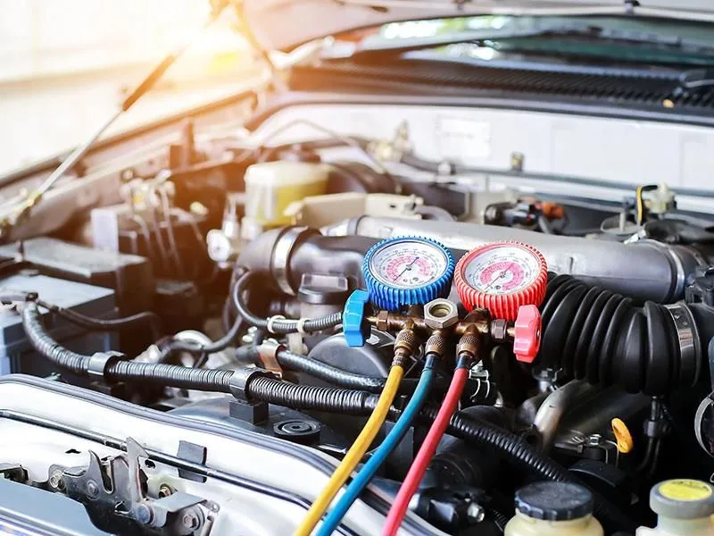 Nahaufnahme eines Automotors mit angeschlossenen farbigen Manometern und Schläuchen zur Druckmessung, Motorraum mit geöffnetem Motorhaube und Sonnenlicht im Hintergrund.