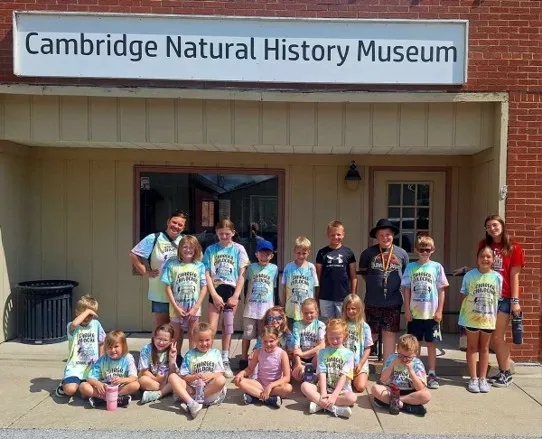 Group of children and two adults wearing bright t-shirts posing in front of the Cambridge Natural History Museum entrance on a sunny day.