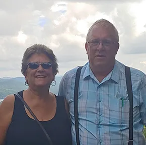 Two people outdoors with cloudy sky and mountains in the background, one wearing sunglasses and the other plaid shirt.