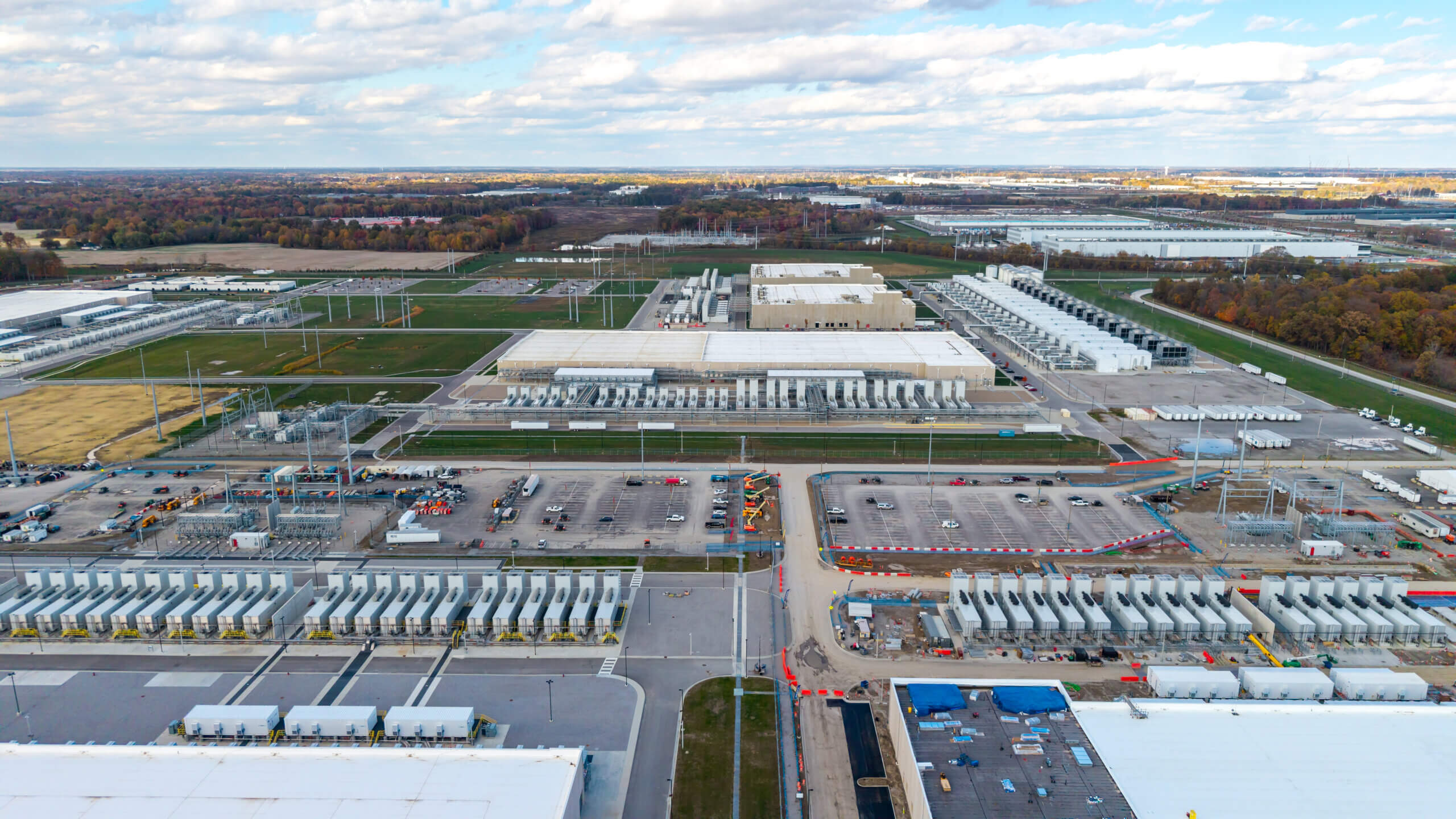 Aerial view of a large data center complex with multiple long, white-roofed buildings, cooling units, parking lots, and electrical infrastructure surrounded by trees and fields under a partly cloudy sky.