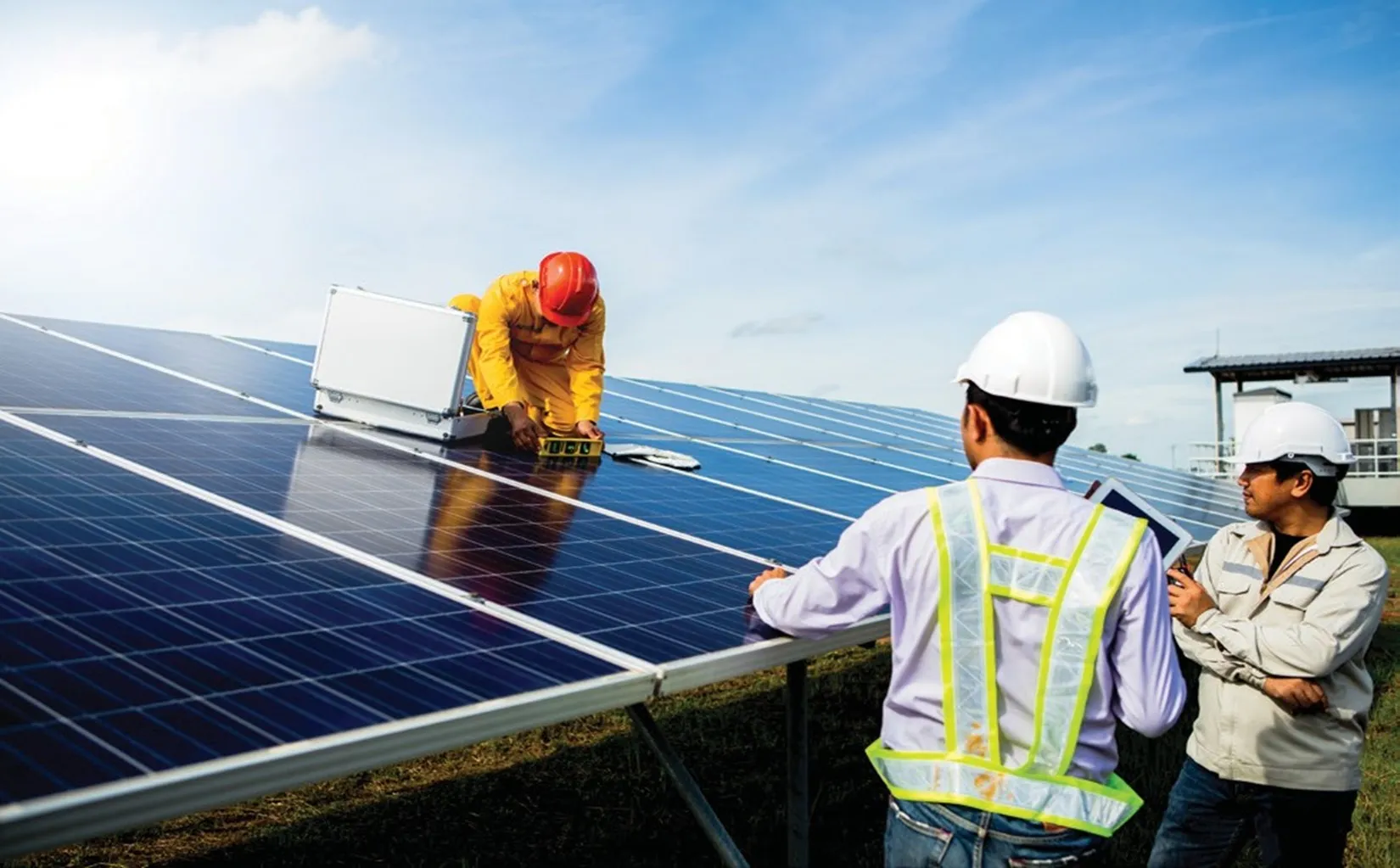 Technicians wearing safety helmets and reflective vests inspecting and working on solar panels under a clear blue sky during daytime.