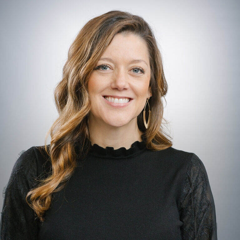 Smiling woman with long wavy brown hair wearing a black top and gold hoop earrings, posed against a light gray background.