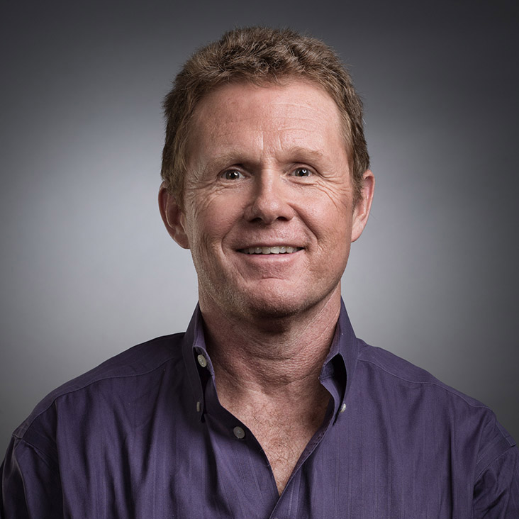 Smiling middle-aged man with light brown curly hair wearing a dark purple button-up shirt against a plain gray background.