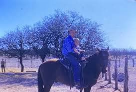 An elderly man in a blue shirt and a young child riding a dark horse together in an open field with bare trees and a clear sky.