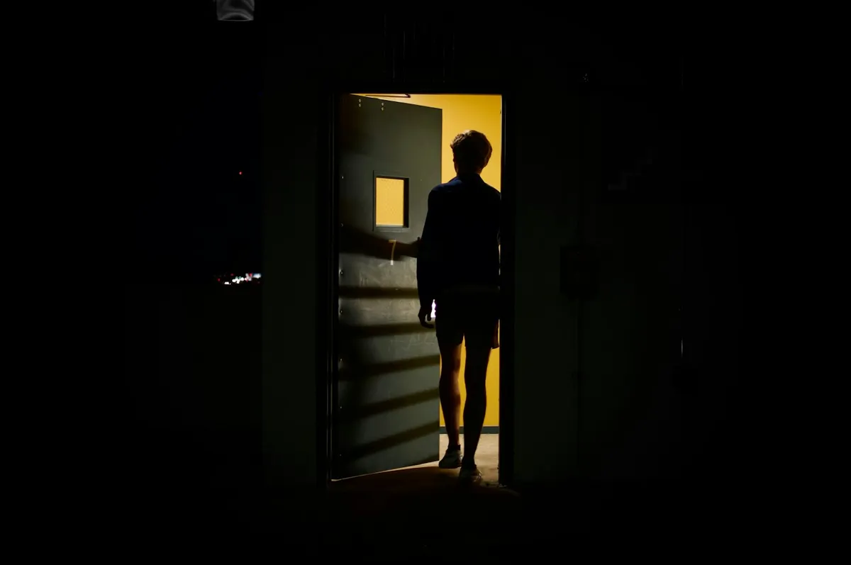 A figure standing in a lit doorway, seen from inside a dark room
