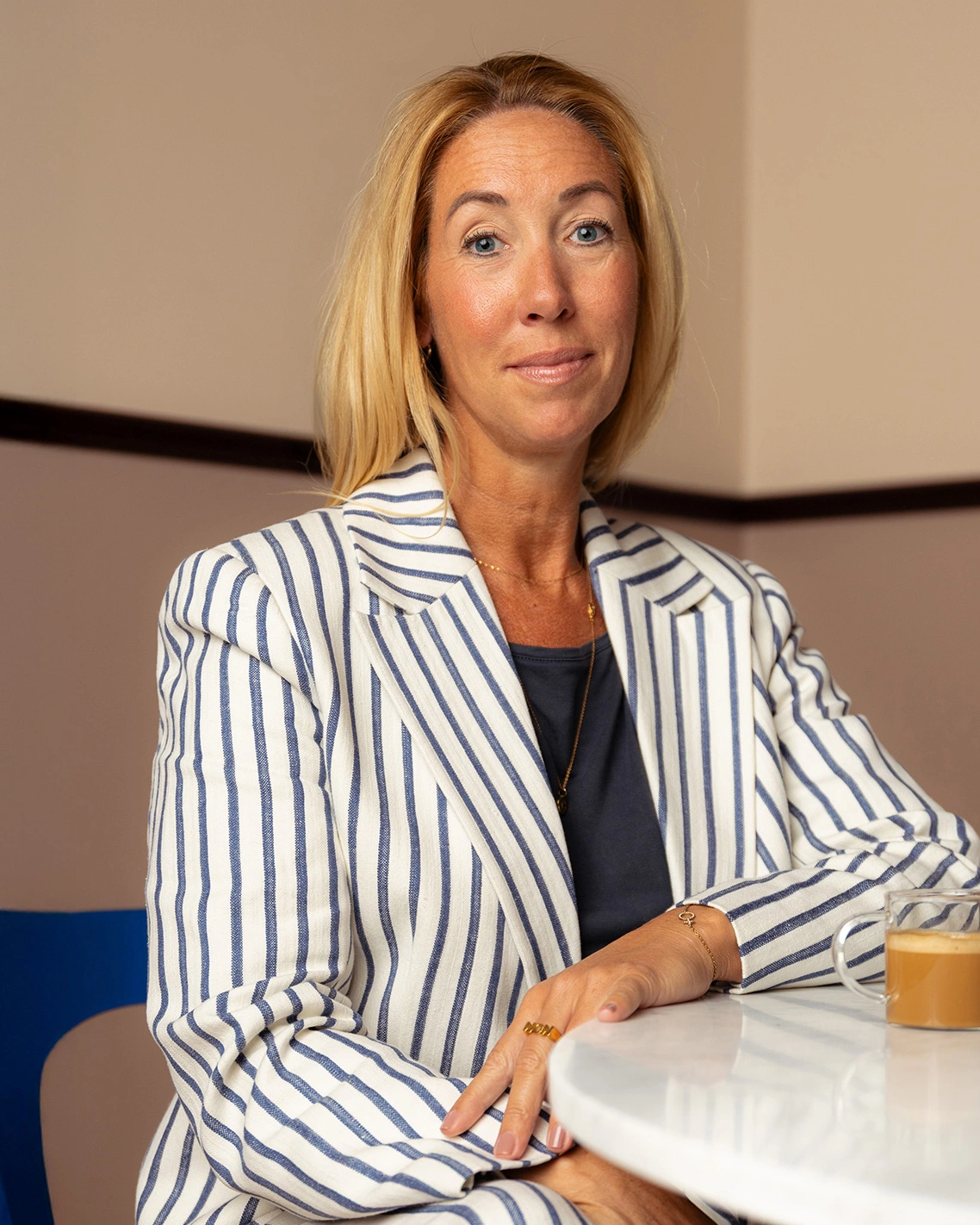Jessica Sparrfeldt trägt einen weißen Blazer mit blauen vertikalen Streifen, sitzt an einem runden weißen Tisch mit einer Kaffeetasse aus klarem Glas und blickt selbstbewusst in die Kamera.