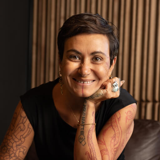 Lächelnde Person mit kurzem dunklem Haar und sichtbaren Tattoos an Armen und Gesicht, bekleidet mit einem ärmellosen schwarzen Oberteil, das Kinn auf die Hand mit einem großen Ring gestützt, sitzend vor vertikalen Holzpaneelen.