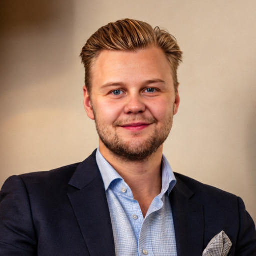 A smiling young man with light hair, blue eyes, and light facial hair, wearing a dark suit jacket over a light blue checked dress shirt, posed against a blurred beige background.