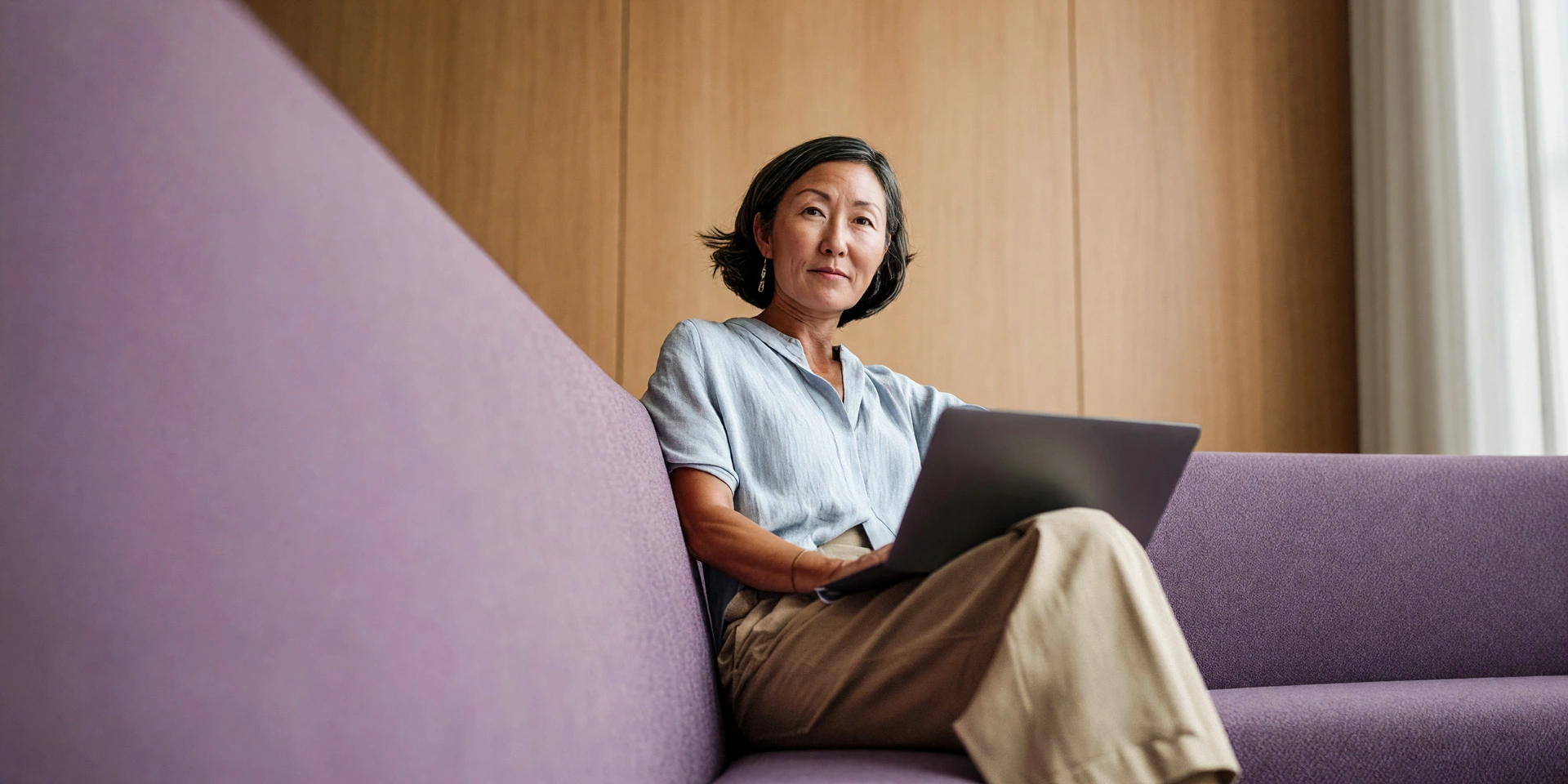 Frau mit kurzem dunklem Haar sitzt auf einem violetten Sofa und arbeitet an einem Laptop; im Hintergrund eine Wand aus Holzpaneelen und ein weißer Vorhang.