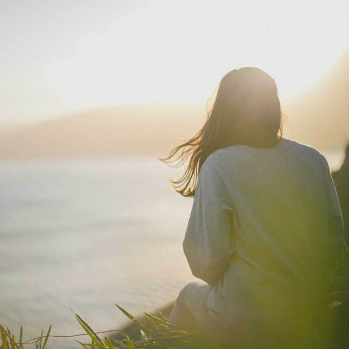 A woman looks pensively into the distance, sat on a hill overlooking. misty, morning seaside. What could she be thinking about?