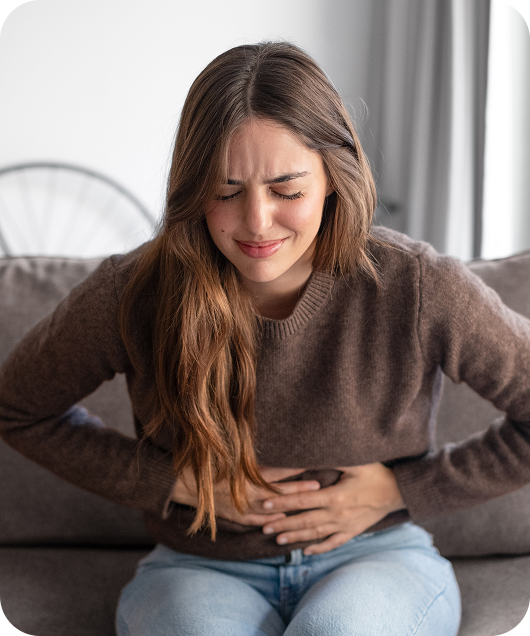 Young woman sitting on a couch with long brown hair wearing a brown sweater and light blue jeans, holding her stomach with a pained expression, indicating discomfort or stomach ache.