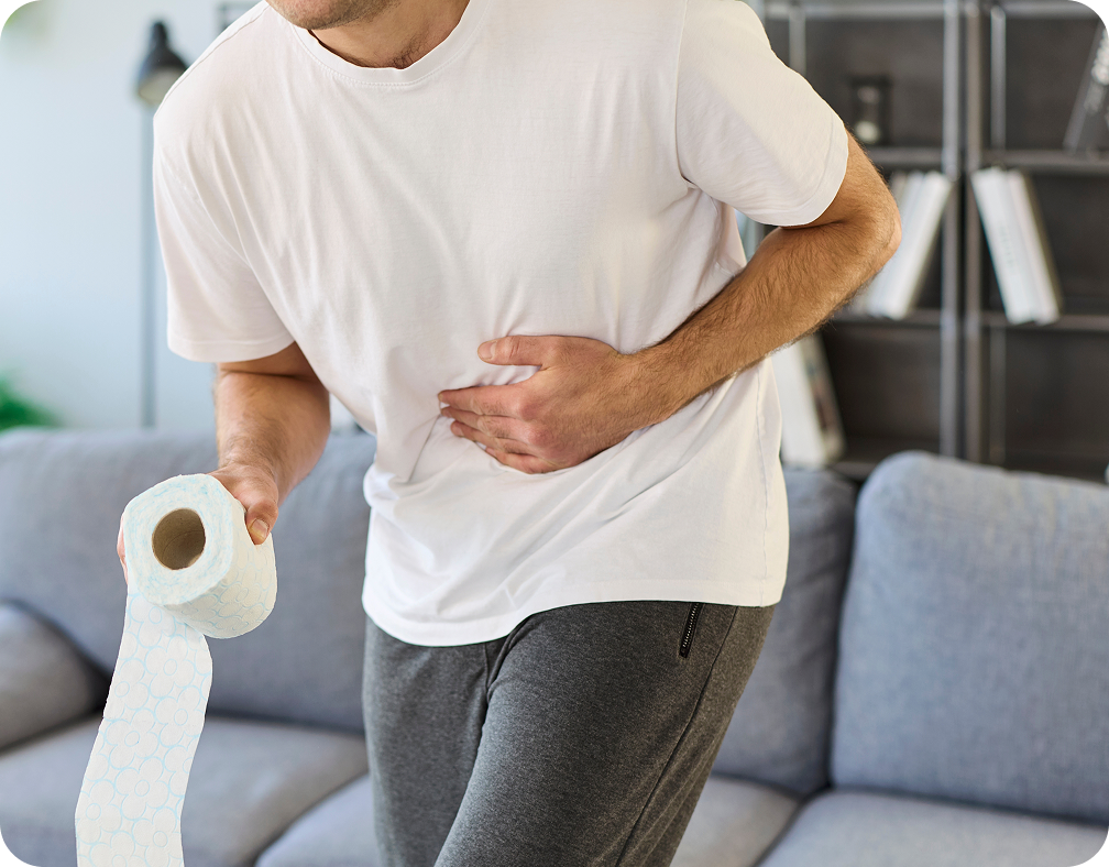 Man in white t-shirt and gray pants holding a roll of toilet paper in one hand and clutching his stomach with the other, appearing to have abdominal pain, standing in a living room with a gray couch and shelves with books in the background.