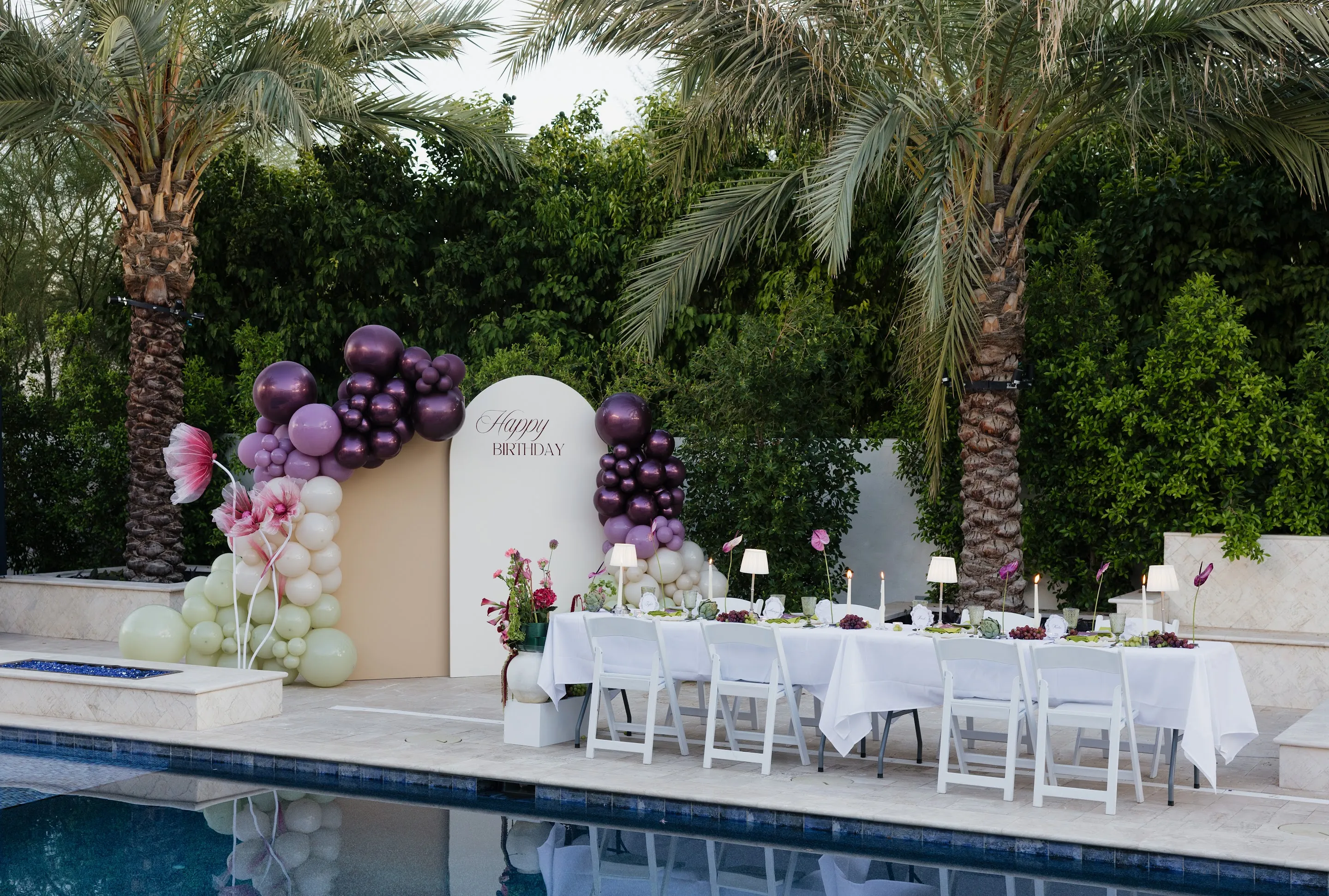 A long white dinner table beside a pool with a plum balloon arch and palm trees