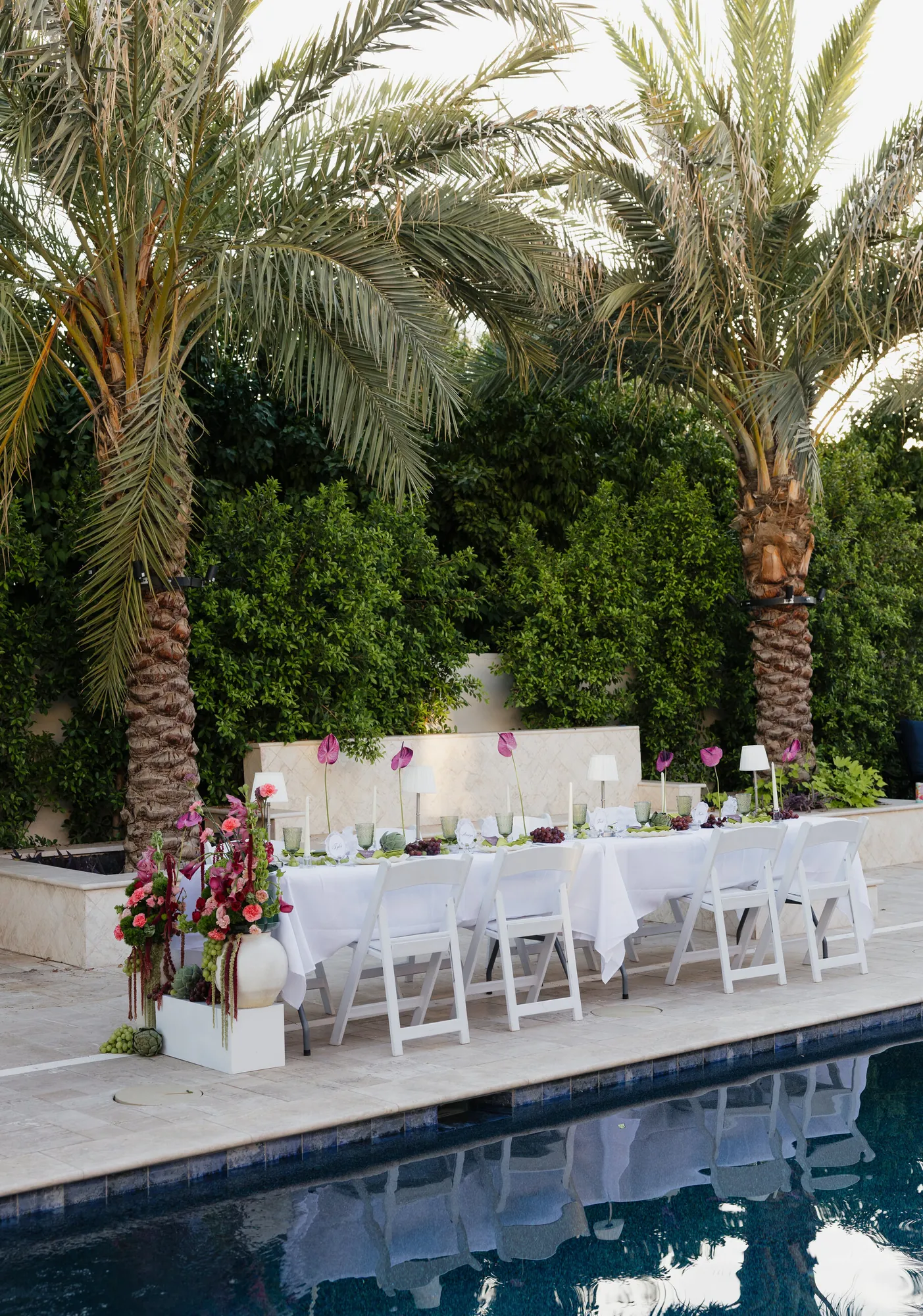 Long dinner table set beside a pool under palm trees