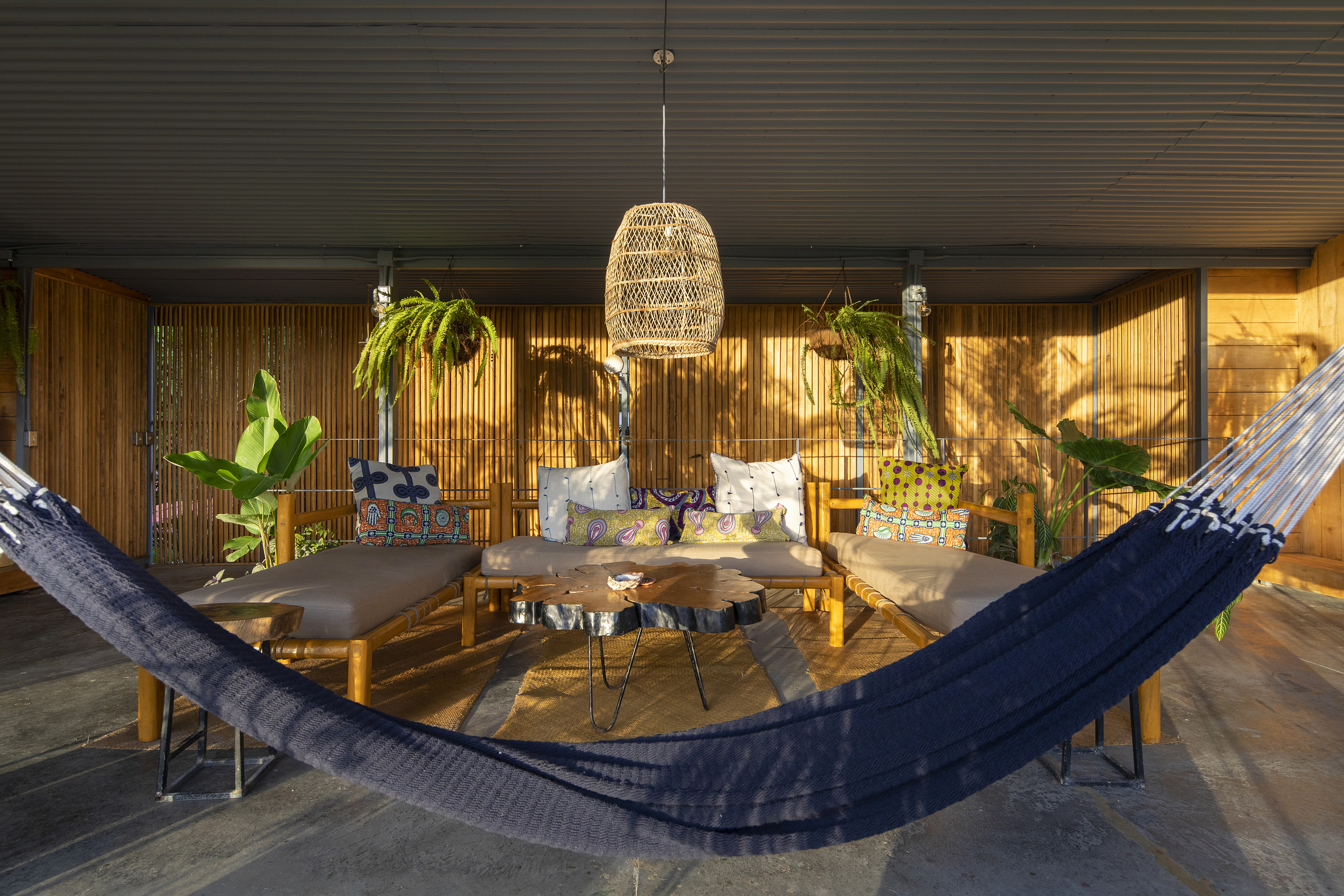 Outdoor seating area with three wooden sofas with beige cushions and colorful patterned throw pillows arranged around a natural wood coffee table, two hanging green ferns, potted plants, a woven ceiling lamp, and a dark blue hammock in the foreground.