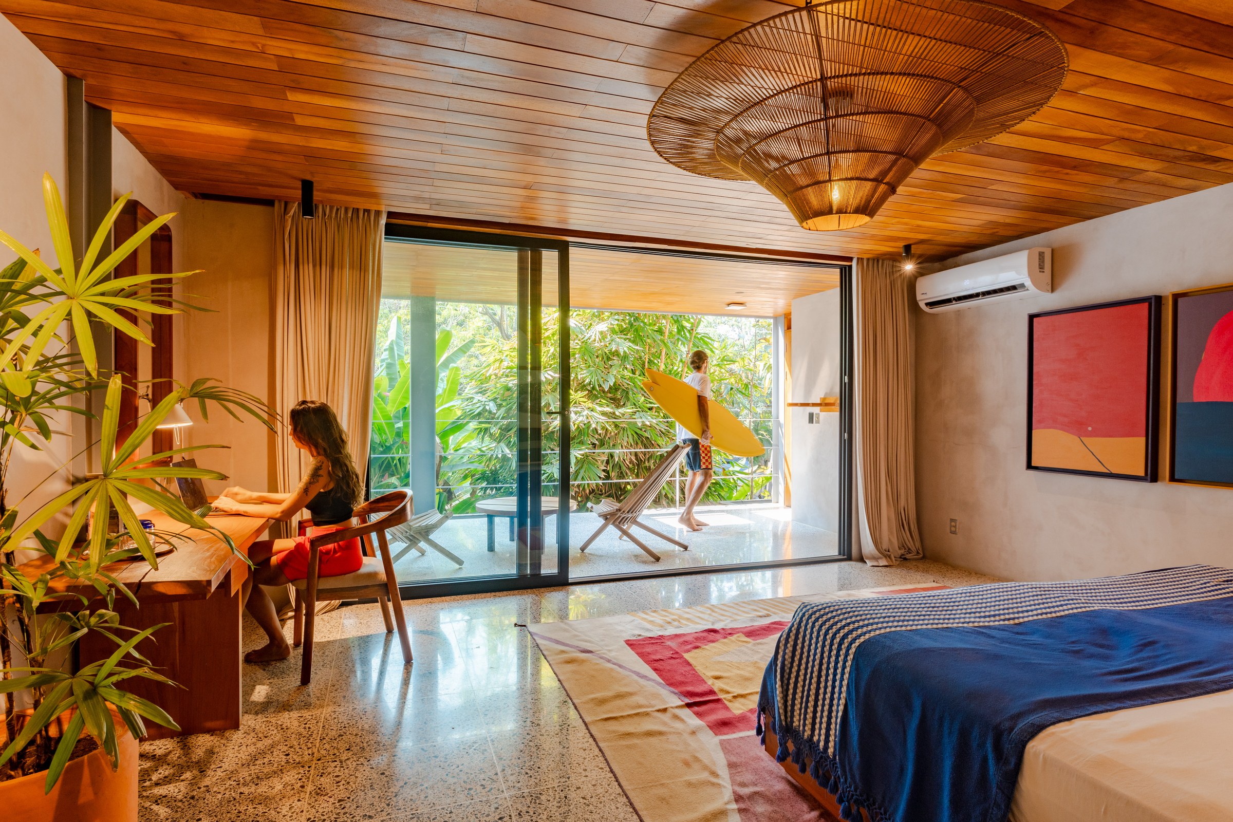 Modern bedroom with a wooden ceiling, large sliding glass doors opening to a balcony surrounded by lush greenery, a woman working on a laptop at a wooden desk, and a man holding a yellow surfboard walking outside.