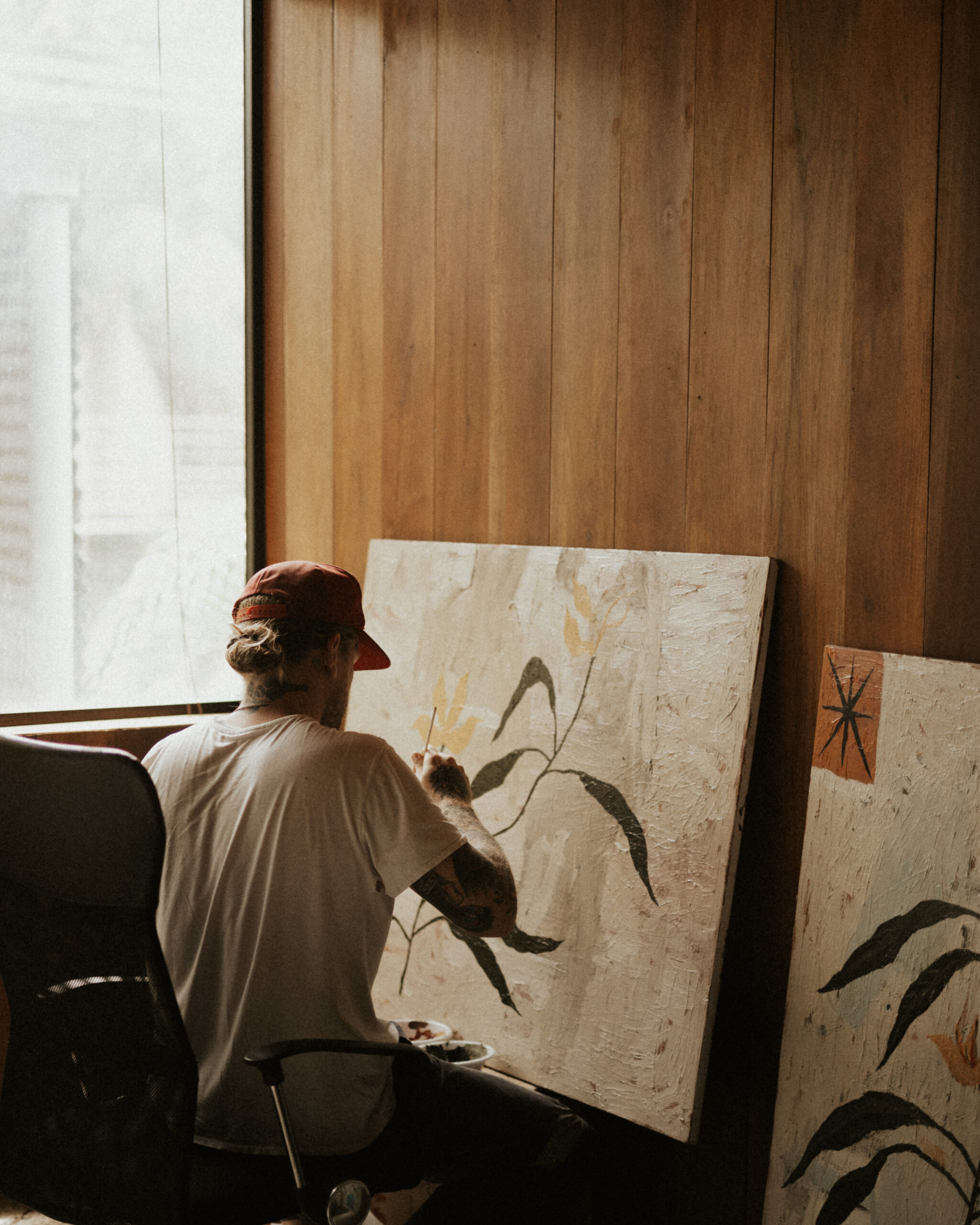 Artist wearing a red cap and white shirt sitting on a black office chair by a large window, painting a canvas with abstract plant designs against a wooden wall.