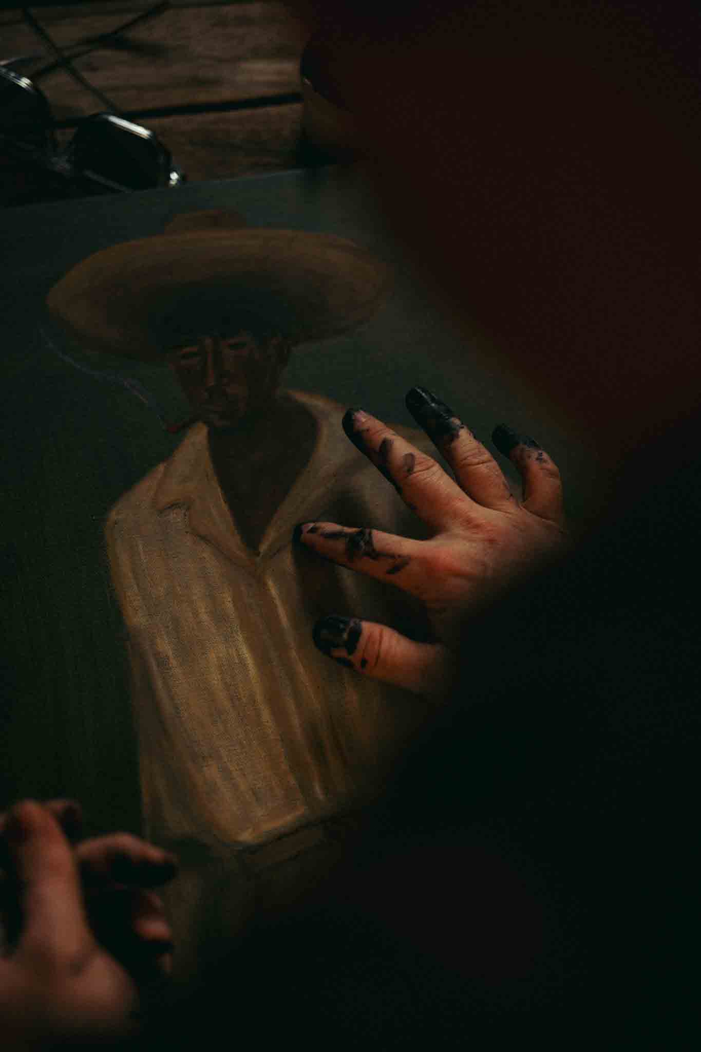 Close-up of a person with paint-covered fingers working on a painting of a man wearing a wide-brimmed hat and light-colored shirt.