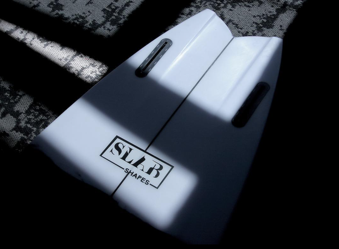 Close-up of a white surfboard with two black fin slots and the logo 'SLAB SHAPES' in black, partially shaded by window shadows.