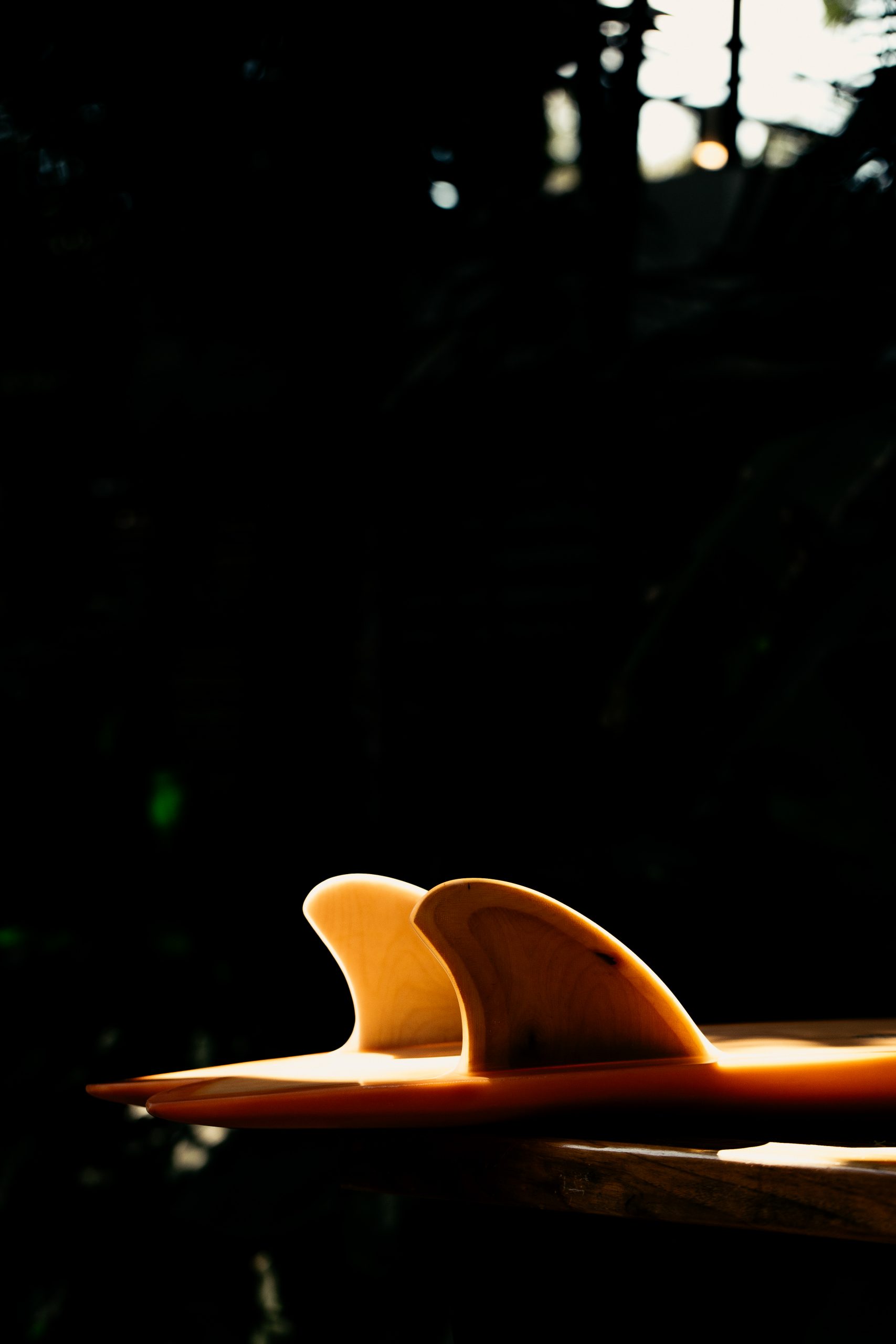 Close-up of two wooden surfboard fins attached to a surfboard, illuminated against a dark background.