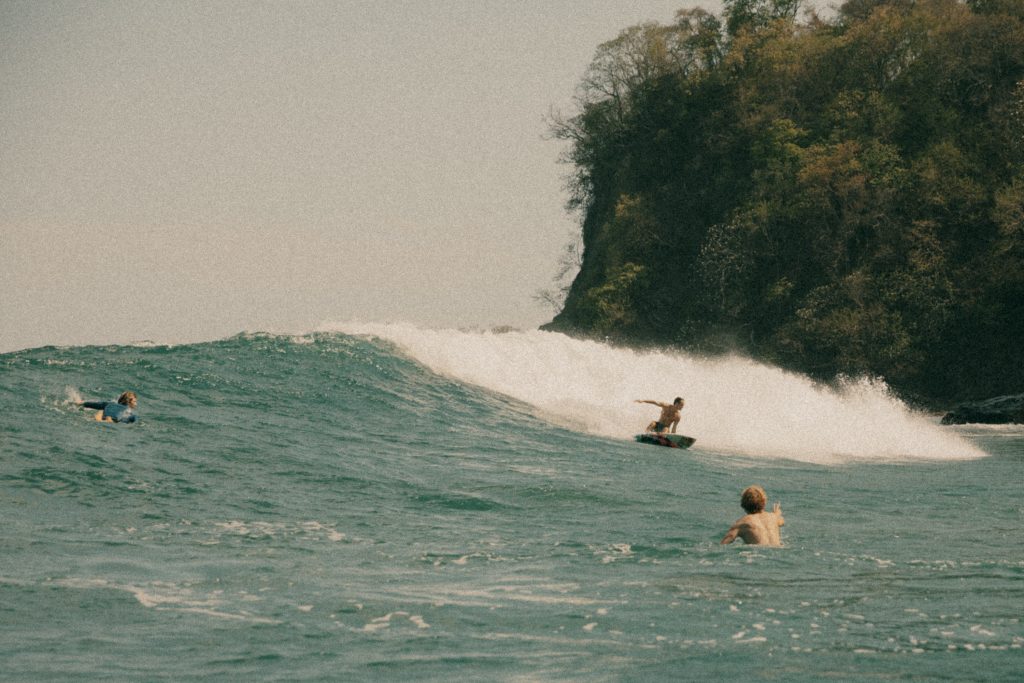 Surfer riding a wave near a forested rocky coastline with two swimmers in the ocean watching.