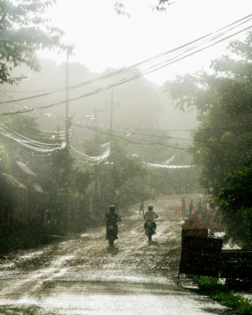 Two motorcyclists wearing helmets ride on a wet, muddy road during rain with blurred trees and power lines overhead.