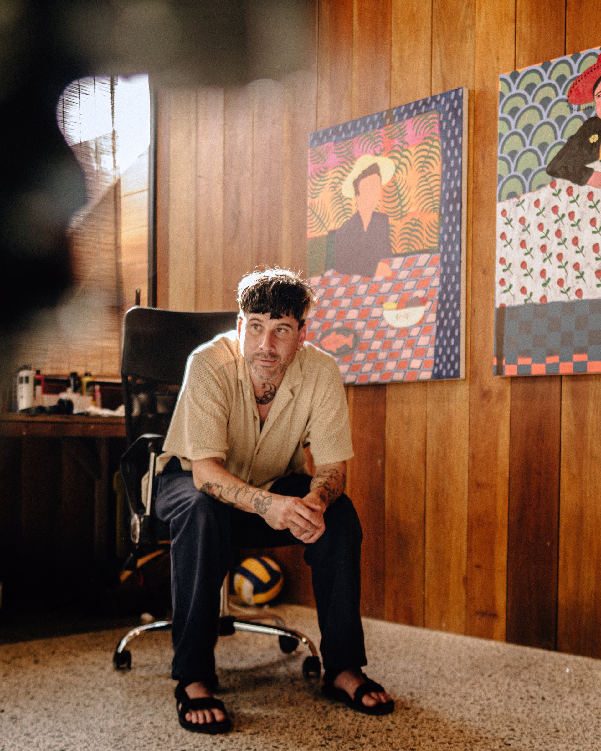 Man with tattoos wearing a beige short-sleeve shirt and black pants sitting on a black office chair in a room with wooden walls, near colorful paintings and a volleyball on the floor.