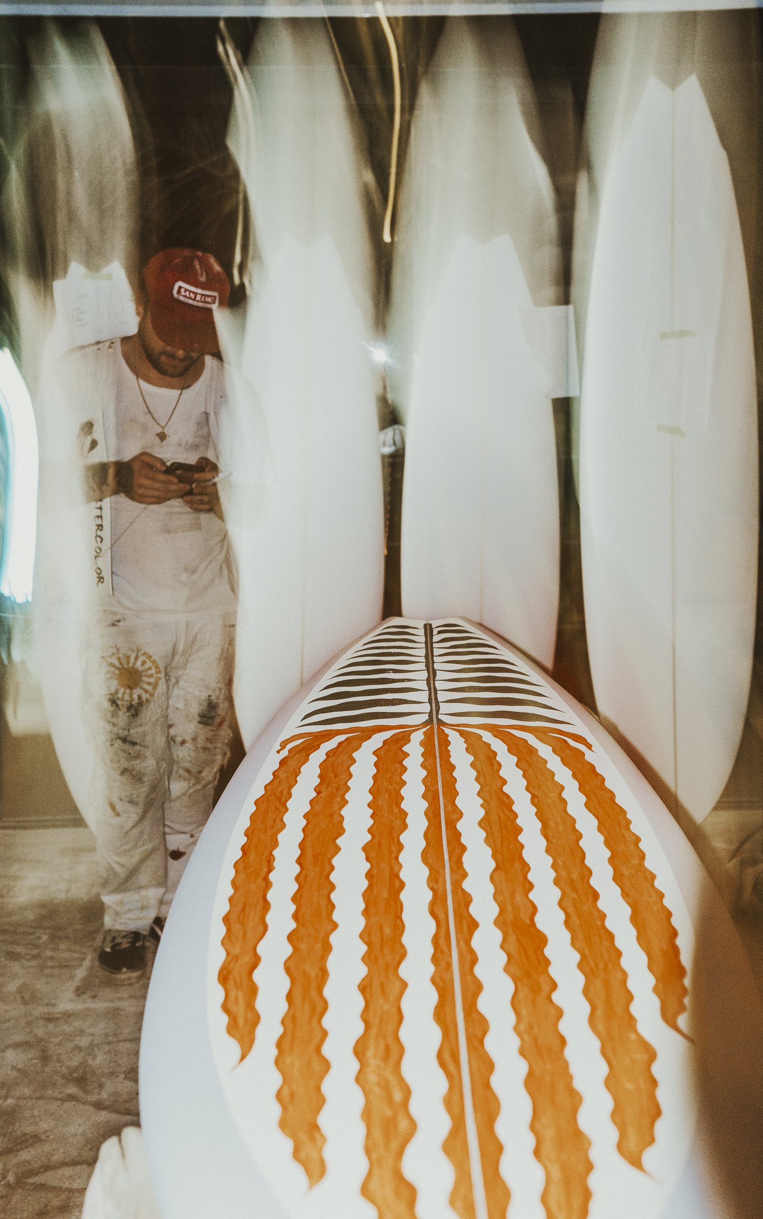 Person wearing a red hat and paint-splattered clothes looking at a phone, standing near surfboards, with a close-up view of a white surfboard featuring orange textured stripes.