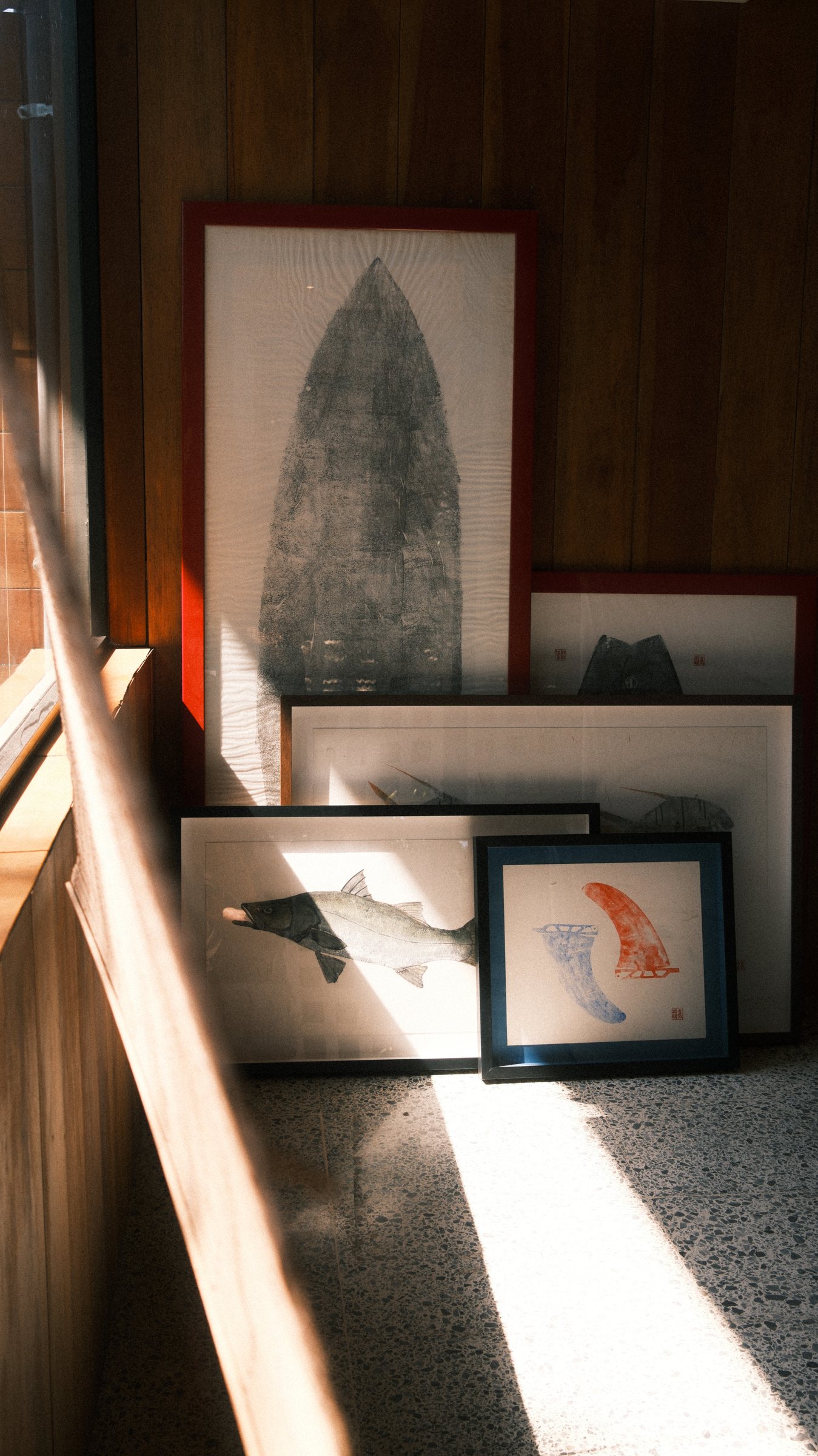 Several framed artworks leaning against a wooden wall, illuminated by sunlight: a large piece with a dark abstract shape in a red frame, a smaller frame with a fish illustration, and another with two colored fins in blue and red.