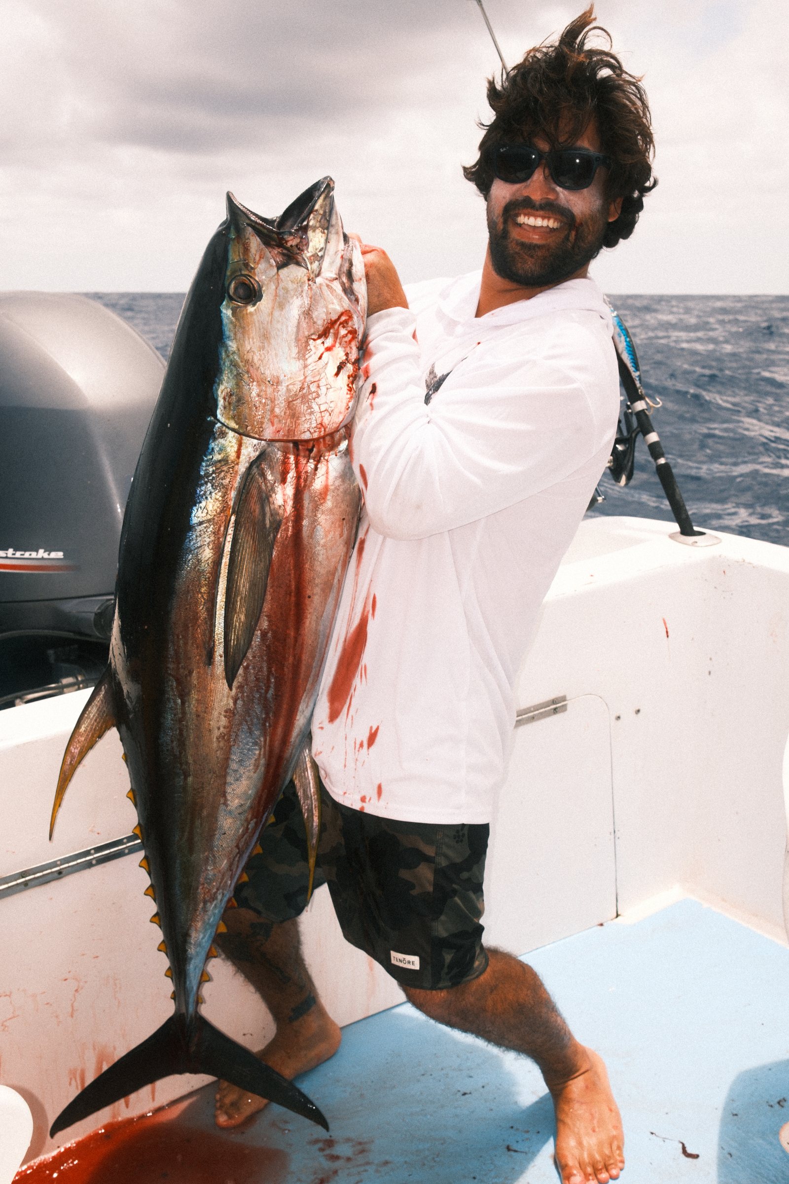 Man with messy hair and sunglasses holding a large bloodied tuna fish on a boat with ocean and cloudy sky in the background.
