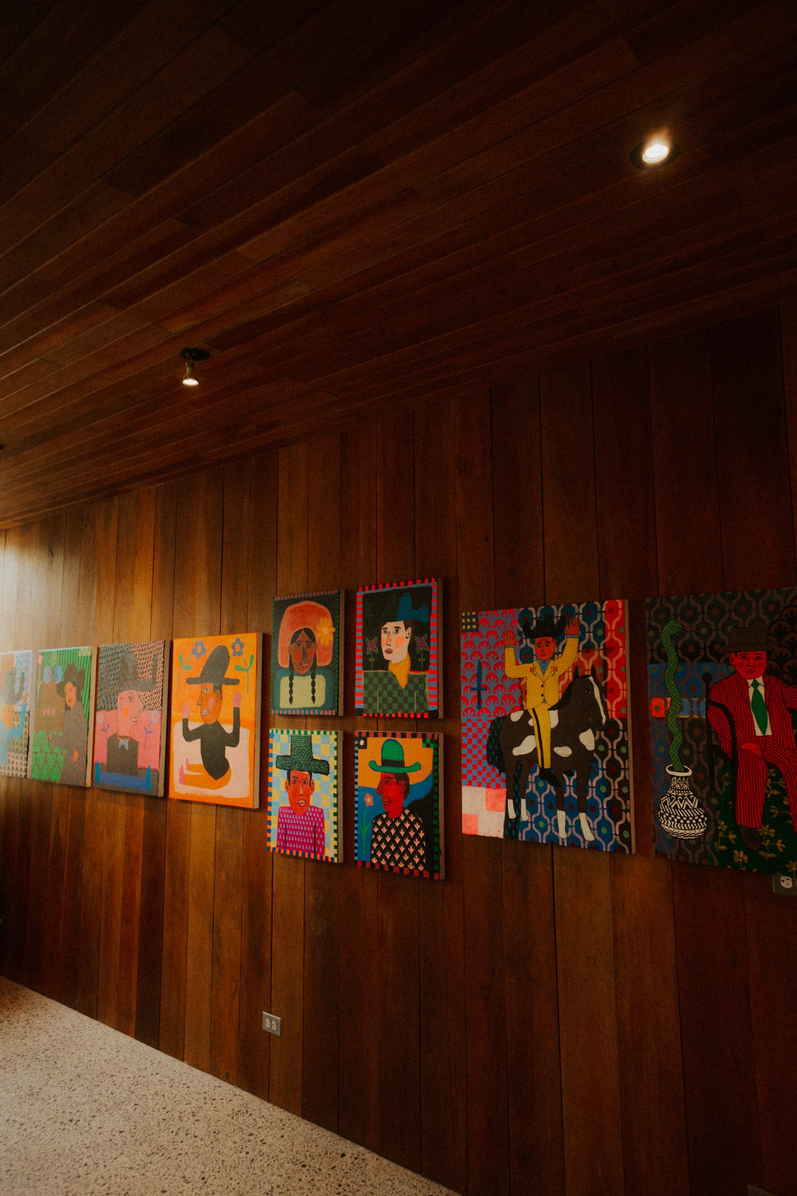 A row of colorful paintings depicting stylized portraits of people wearing hats, displayed on a wooden wall in a warmly lit indoor space.
