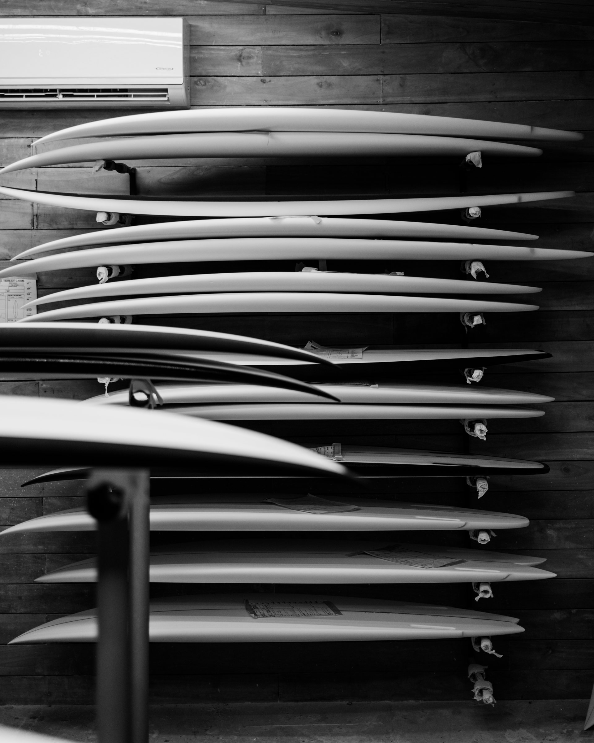 Black and white photo of multiple surfboards stacked horizontally on a wooden wall rack indoors, with an air conditioning unit mounted above.
