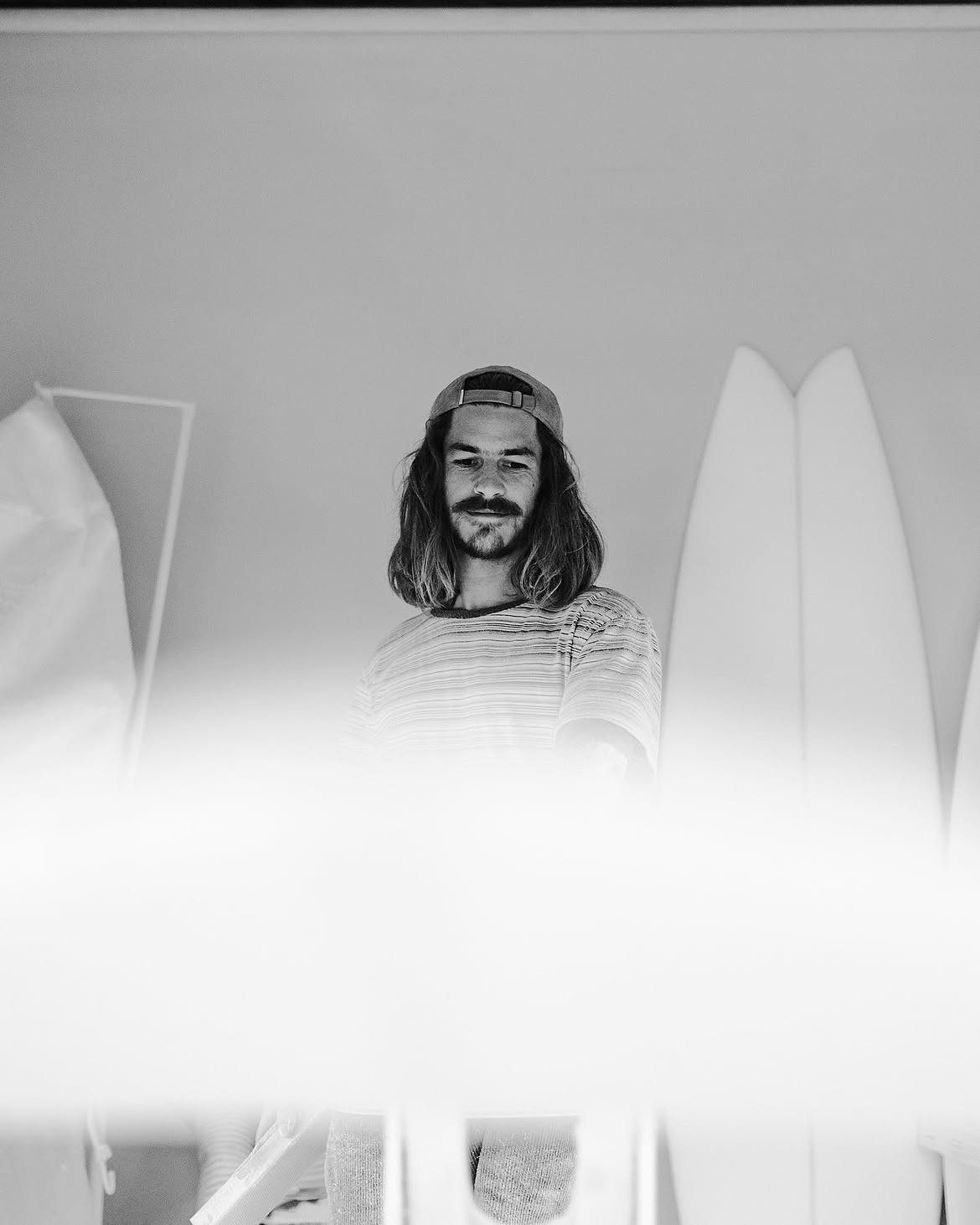 Black and white photo of a man with shoulder-length hair and a beard wearing a backwards cap and striped shirt, standing in front of surfboards indoors, looking downwards with a slight smile.