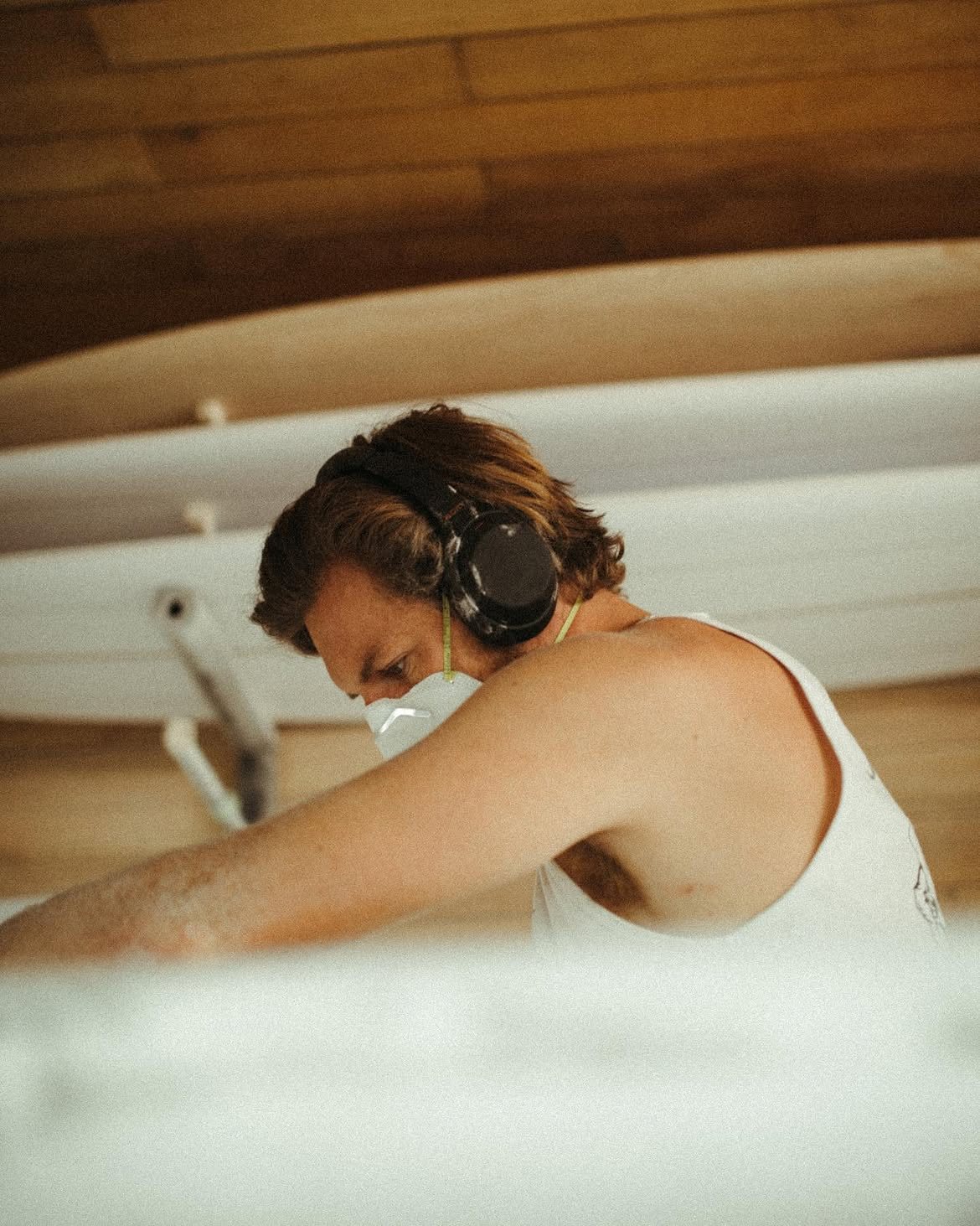 A man wearing a sleeveless white shirt, black headphones, and a protective face mask is working intently, possibly shaping or sanding a long white object under a wooden ceiling.