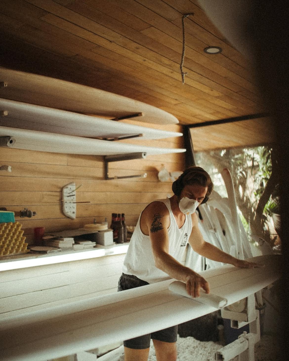 A man wearing a white sleeveless shirt with a tattoo on his upper arm and a protective mask uses a jigsaw to cut wood indoors, with sawdust flying.