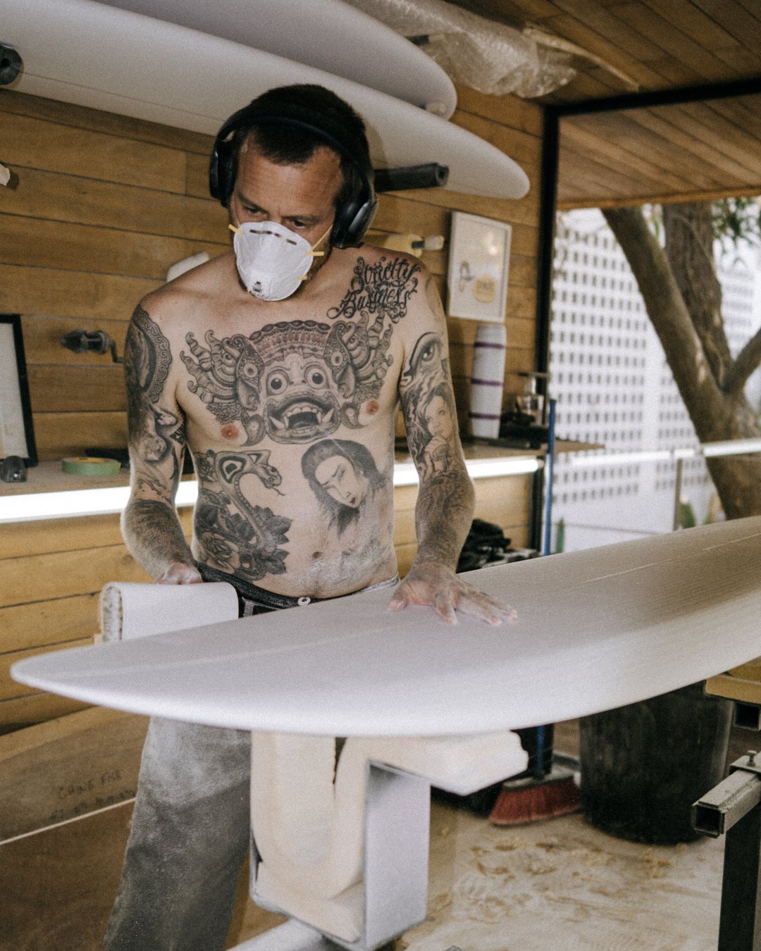 Shirtless tattooed man wearing a protective mask and headphones sanding a white surfboard in a wooden workshop.