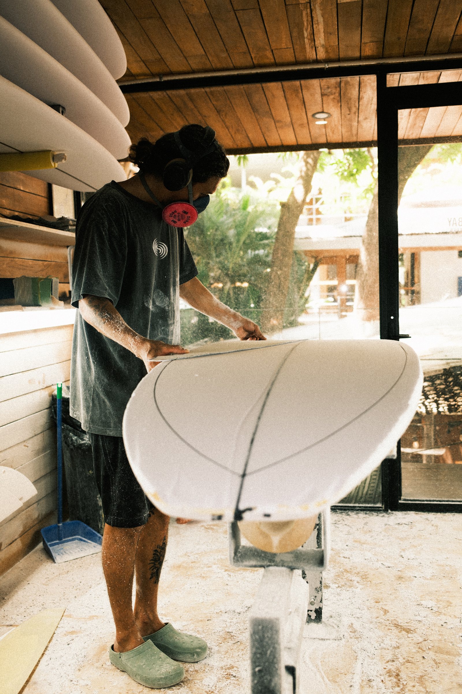 Person wearing a black shirt, black shorts, green slip-on shoes, headphones, and a pink respiratory mask, shaping a white surfboard inside a workshop with wooden walls and multiple surfboards stacked on the side.