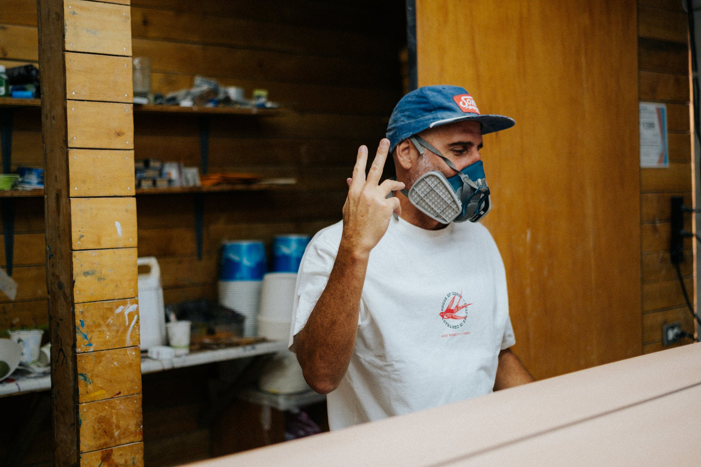 A masked board builder in a blue cap works in a wood-lined workshop.